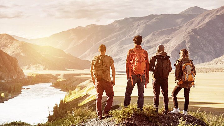 Piękny, ciepły dzień. Grupa młodych osób z turystycznymi plecakami stoi zwrócona tyłem do obserwatora i podziwia oświetlone słońcem góry. 